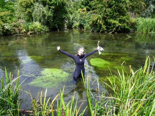 Kathy Hughes in a wetsuit, standing with her arms raised in a pool with shrubs behind her and reeds in front