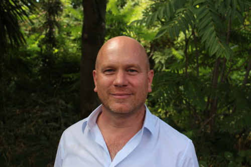Brian Zimmerman, wearing an open-necked shirt, with dense vegetation behind him, smiling at the Camera