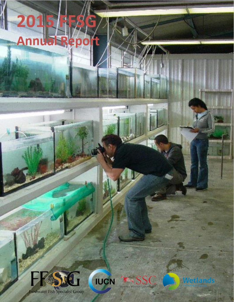 Front cover of the FFSG 2015 Annuarl Report - Three shelves of fish tanks in a steel building with a concrete floor. Three people are present - two men are taking photographs of the fish in the tanks, and a woman has a clip board and is writing something.